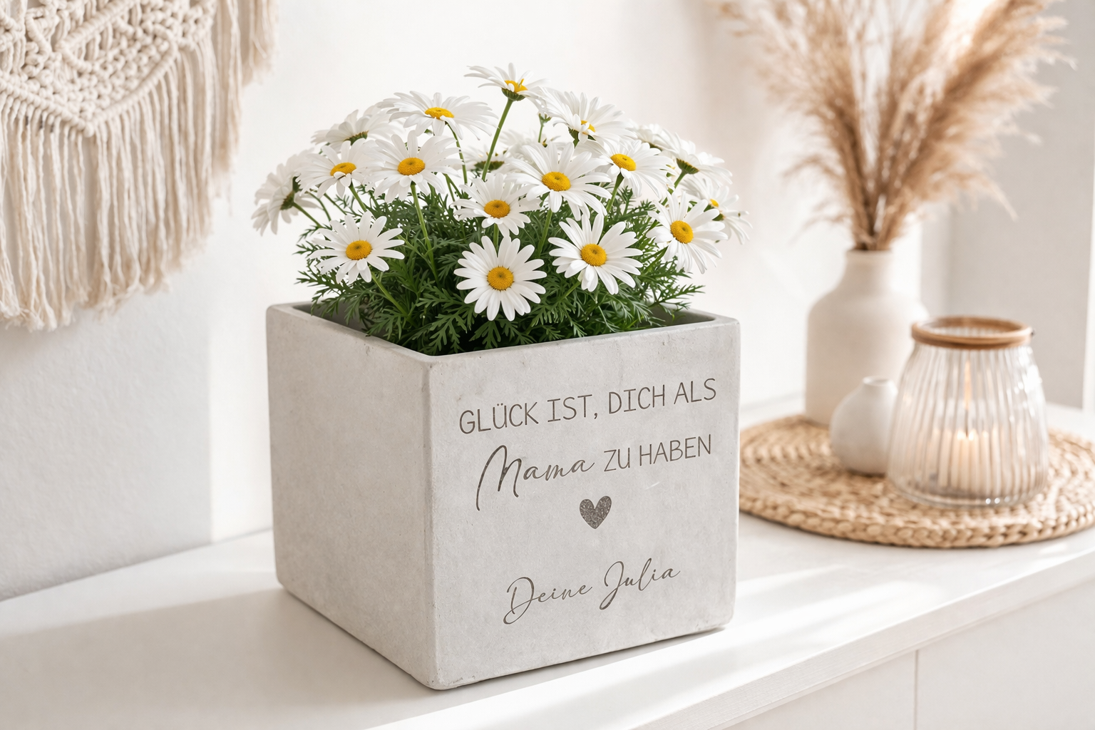 Moderner Blumentopf mit Gravur im Betonlook mit zwei weißen Margeriten Pflanzen. Der personalisierte Blumentopf steht auf einem hellen Sideboard mit Boho Dekoration und Kerzen. Perfektes Muttertagsgeschenk oder Geschenk für Mama und Oma. Ideal für Menschen, die kleine persönliche Geschenke suchen und gerne Kleinigkeiten schenken.