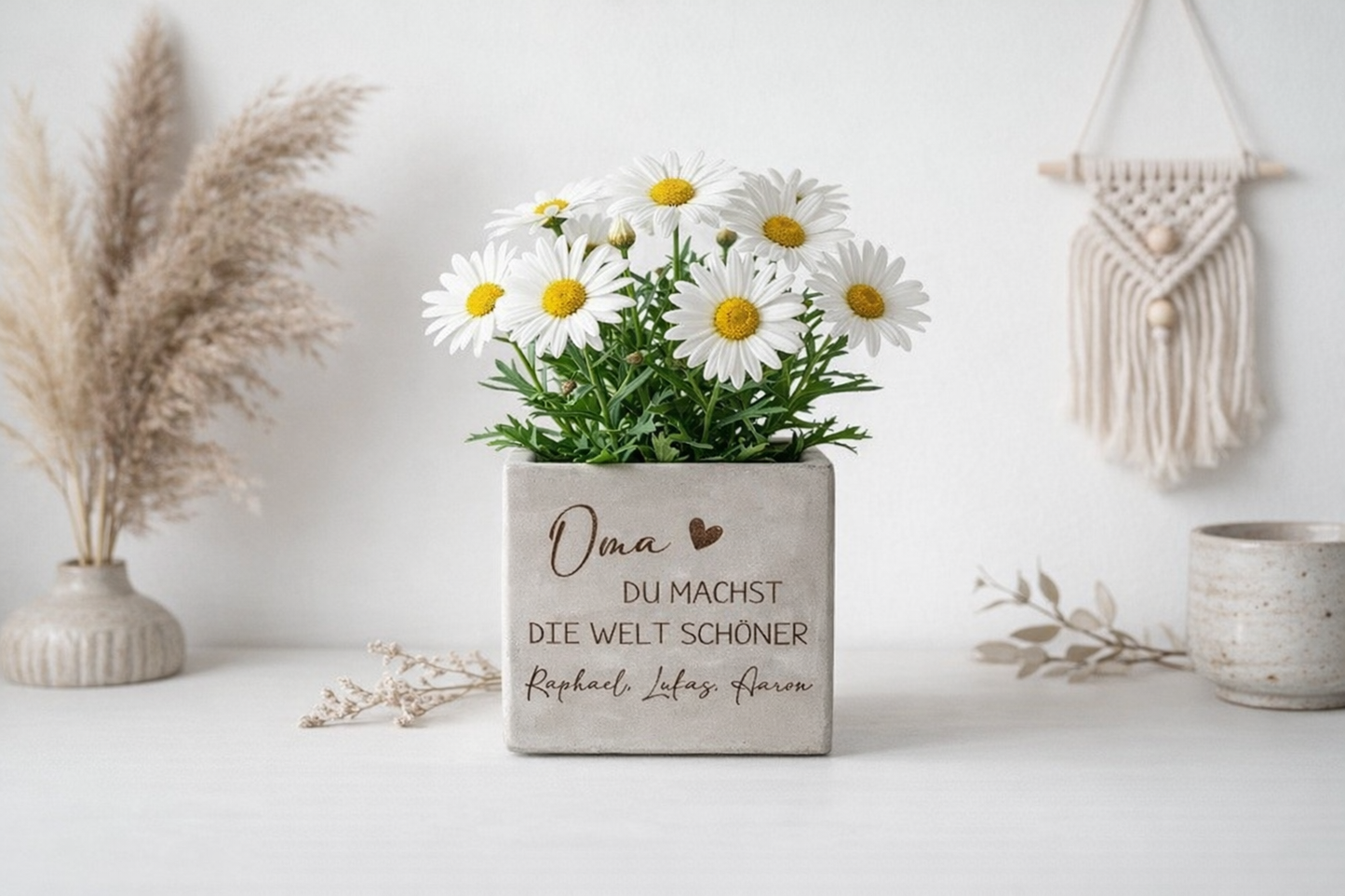 Personalisierter Blumentopf mit Gravur in moderner Betonoptik mit der Aufschrift „Oma – Du machst die Welt schöner“. Dekorativer Blumenübertopf mit weißen Margeriten auf hellem Hintergrund mit Kerzen und Boho Dekoration. Perfektes Muttertagsgeschenk oder liebevolles Geschenk für Oma. Individueller Blumentopf mit Namen der Enkel als personalisierte Geschenkidee zum Geburtstag, Weihnachten oder Ostern für Familie.
