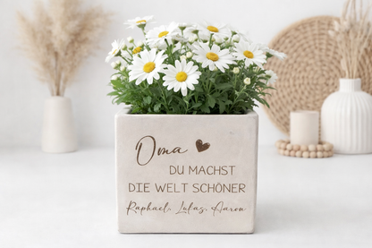 Personalisierter Blumentopf mit Gravur in moderner Betonoptik mit der Aufschrift „Oma – Du machst die Welt schöner“. Dekorativer Blumenübertopf mit weißen Margeriten auf hellem Hintergrund mit Kerzen und Boho Dekoration. Perfektes Muttertagsgeschenk oder liebevolles Geschenk für Oma. Individueller Blumentopf mit Namen der Enkel als personalisierte Geschenkidee zum Geburtstag, Weihnachten oder Ostern für Familie.