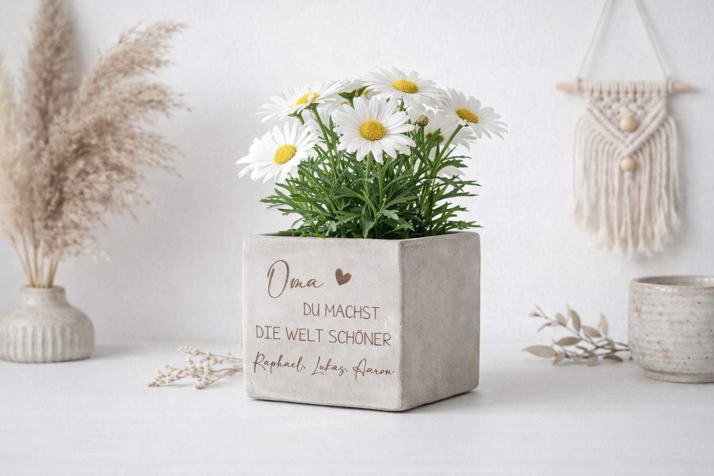 Personalisierter Blumenübertopf mit Gravur und Herzmotiv für Oma oder Mama. Dekorativer Blumentopf in Betonoptik mit Margeriten auf hellem Sideboard mit Boho Dekoration. Emotionales Geschenk für Familie mit individueller Namensgravur. Perfekt als Muttertagsgeschenk, Geburtstagsgeschenk oder kleine Aufmerksamkeit für besondere Menschen.