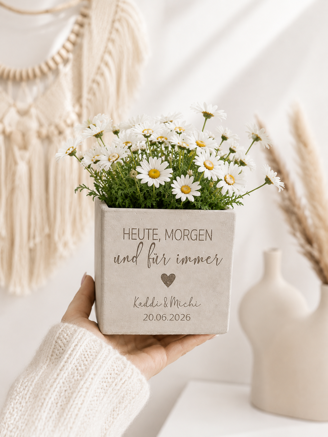 Blumentopf mit Gravur und persönlicher Widmung als stilvolles Hochzeitsgeschenk. Dekoriert mit weißen Margeriten und fotografiert in einer Hand vor modernem Boho-Hintergrund.
