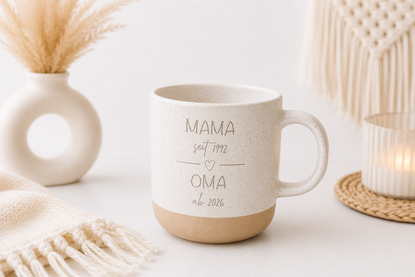 Beige gesprenkelte Keramiktasse mit Gravur „Mama seit 1992 Oma ab 2026“ auf hellweißem modernen Hintergrund mit Makramee, Kerze und Boho Dekoration.