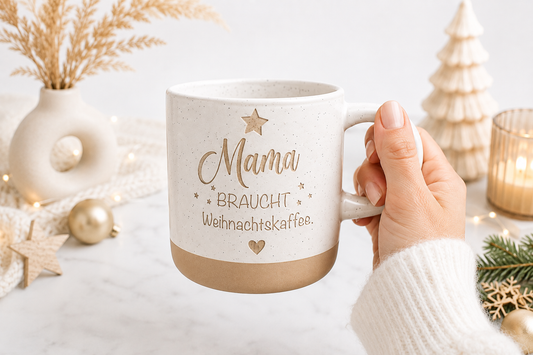 Weiße Steingut Tasse mit Gravur Mama braucht Weihnachtskaffee, gehalten in einer Hand vor heller weihnachtlicher Boho-Dekoration. Liebevolle Geschenkidee für Mama zu Weihnachten.
