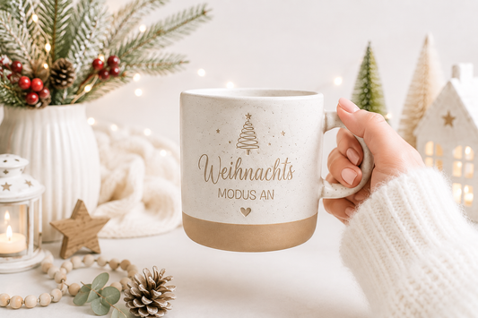 Weihnachtstasse aus Keramik mit Spruch „Weihnachtsmodus an“, liebevoll in einer Hand gehalten. Umgeben von Tannenzweigen und dezenter Weihnachtsdeko. Geschenkidee für Kaffeeliebhaber.